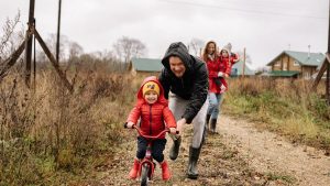 A happy family enjoys a bonding moment outdoors with a child learning to ride a bicycle on a fall day.